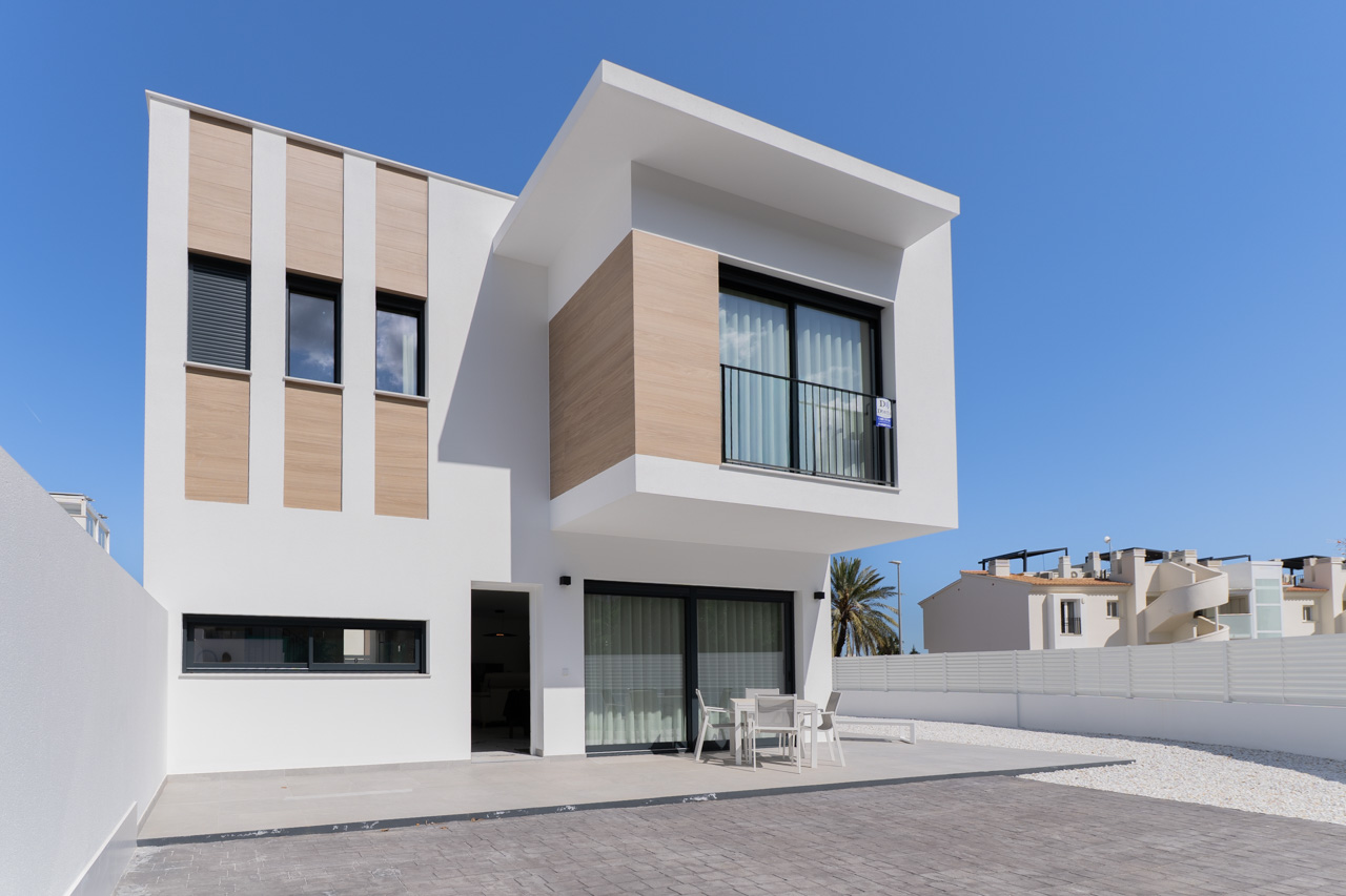 Moderne Außenfassade mit Holzoptik-Paneelen, gepflasterter Terrasse mit Gartenmöbeln und großen Fenstern mit schwarzen Rahmen.