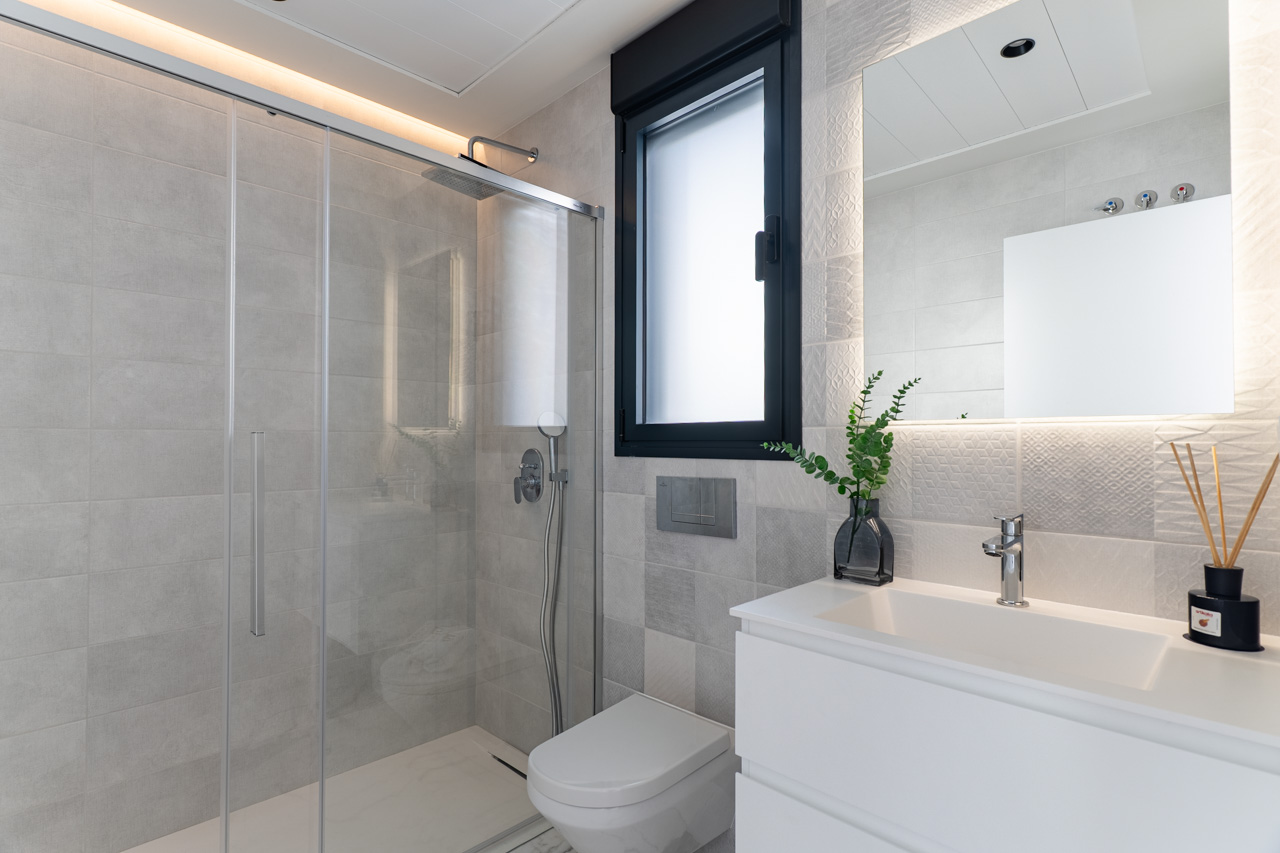 Modernes Badezimmer mit begehbarer Glasdusche, strukturierten grauen Wandfliesen, weißem Waschtisch und einem hintergrundbeleuchteten Spiegel mit LED-Licht.