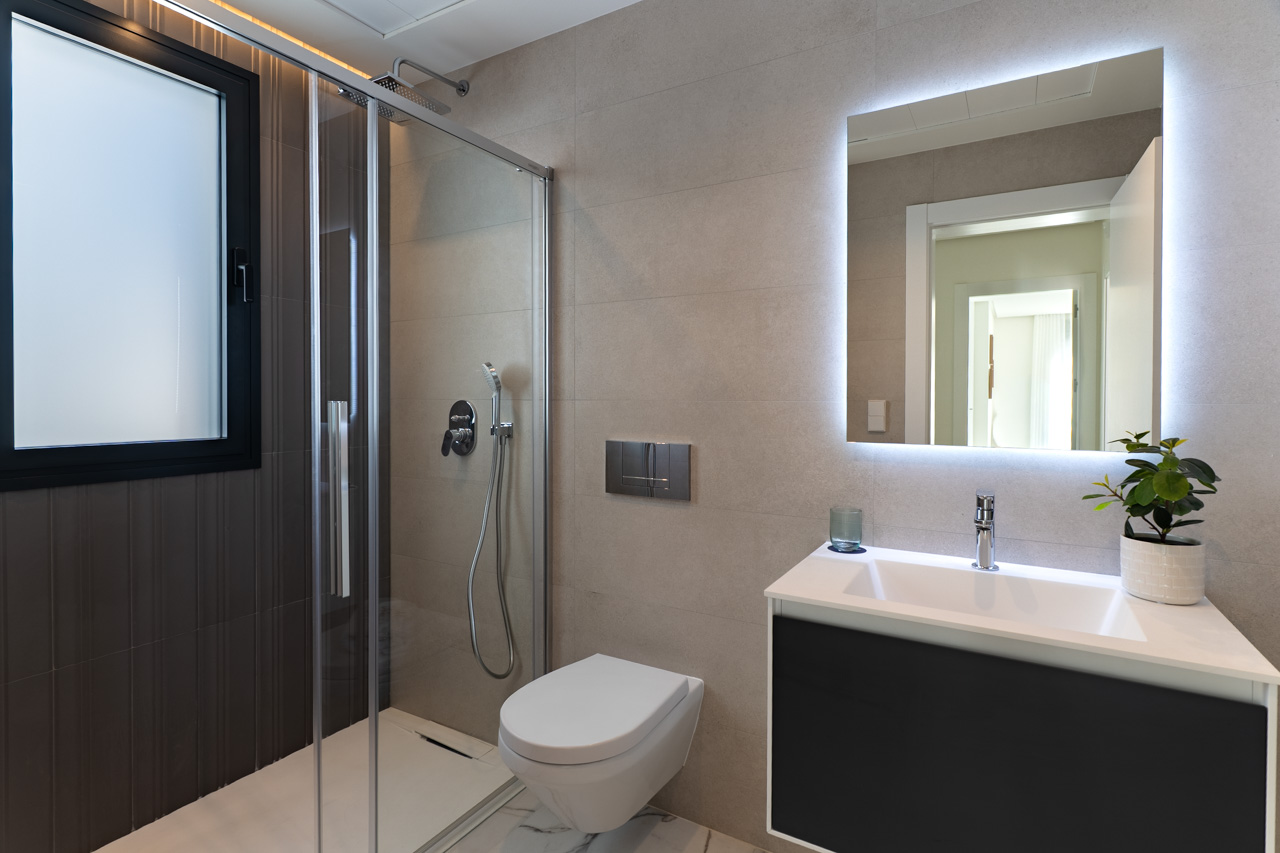 Modernes Badezimmer mit bodengleicher Dusche, Glastrennwand, wandhängendem WC und Waschtisch mit hintergrundbeleuchtetem LED-Spiegel auf beigen Fliesen.