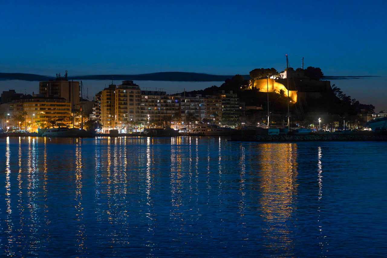 Nachtansicht des Hafens von Dénia mit der beleuchteten Burg auf dem Hügel und den Spiegelungen der Stadtlichter auf dem Mittelmeer.