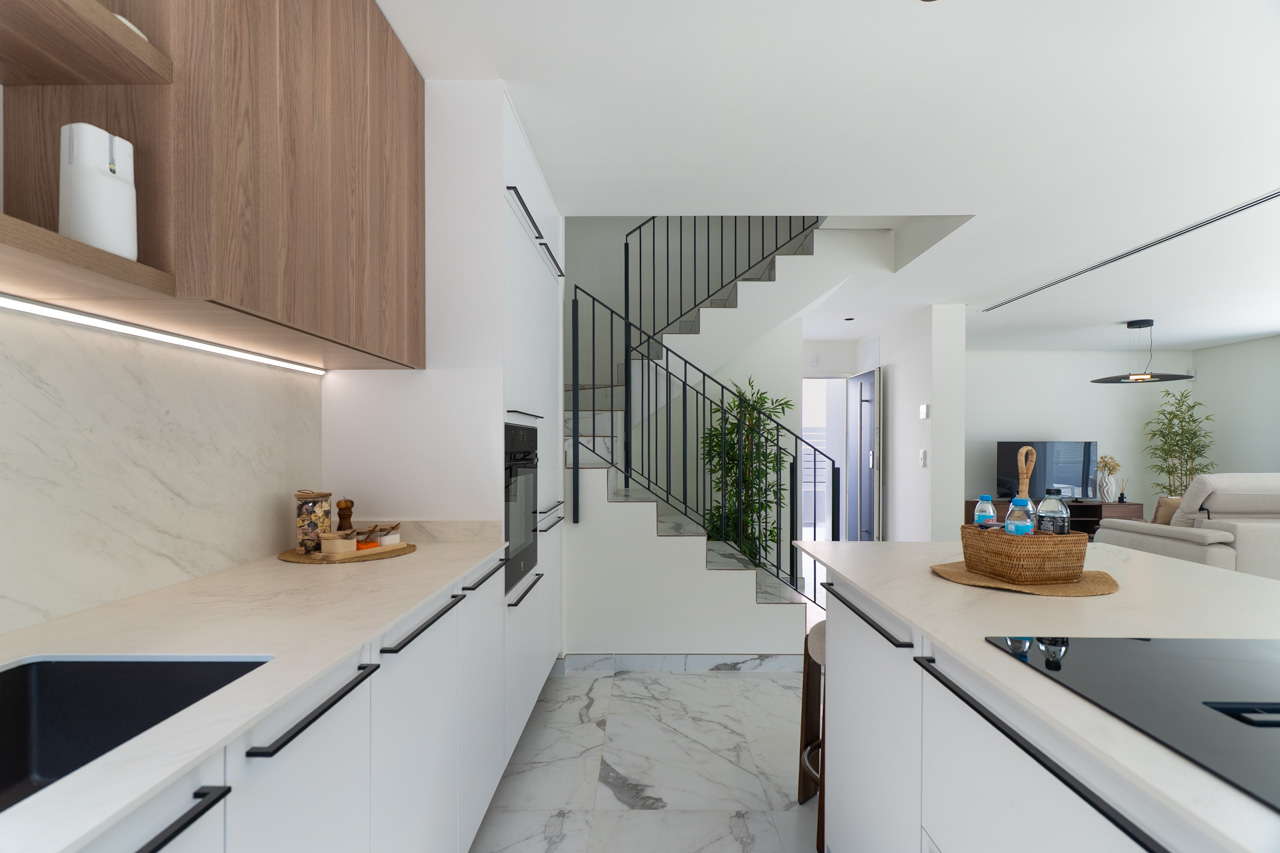Offene Küche mit weißen Fronten, Arbeitsplatten in Marmoroptik und Holzoberschränken mit LED-Beleuchtung. Im Hintergrund sind die Designtreppe und das Wohnzimmer sichtbar.