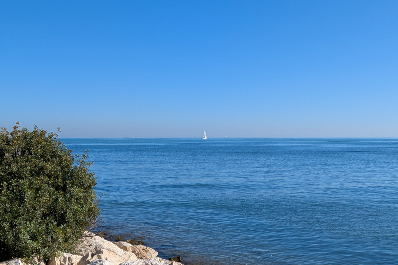 Panoramablick auf das Mittelmeer von der felsigen Küste Dénias aus, mit Segelbooten am Horizont unter strahlend blauem Himmel.