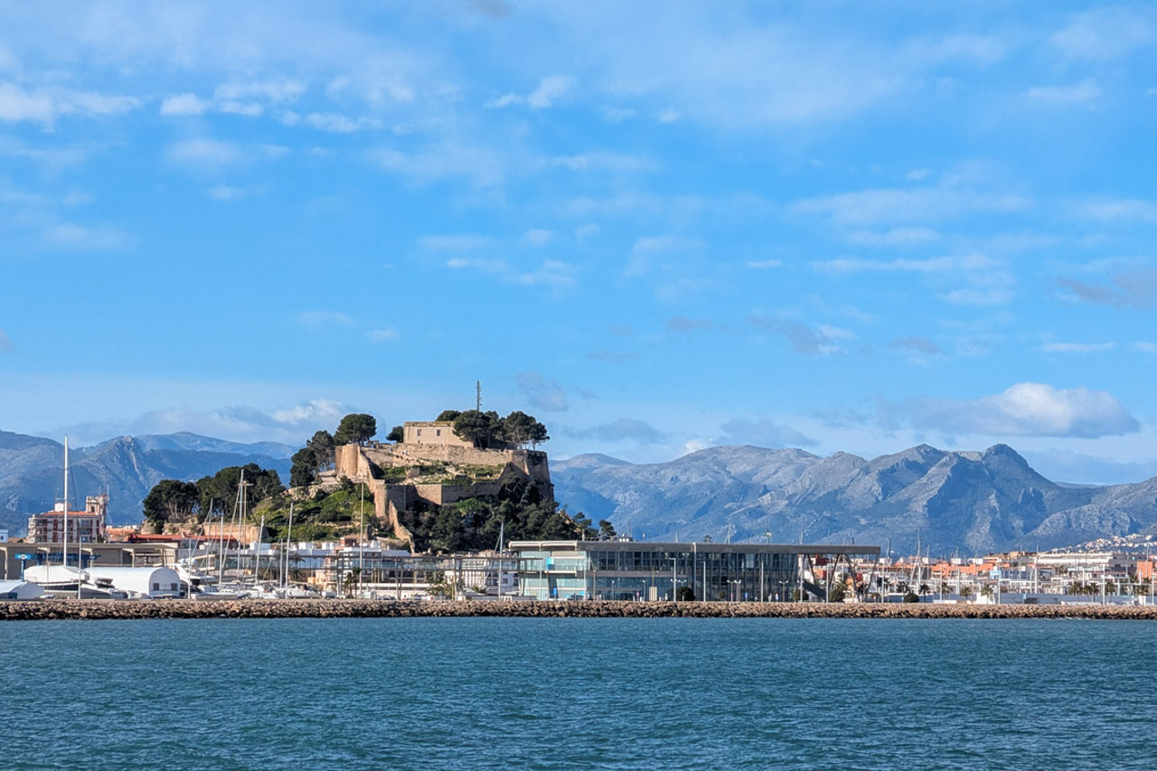 Panoramablick auf den Hafen von Dénia mit der historischen Burg und dem Montgó-Massiv im Hintergrund, gesehen von der Wasserseite aus.
