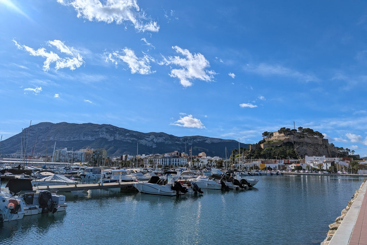Panoramablick auf den Yachthafen von Dénia mit der historischen Burg und dem Montgó-Massiv im Hintergrund unter blauem Himmel.
