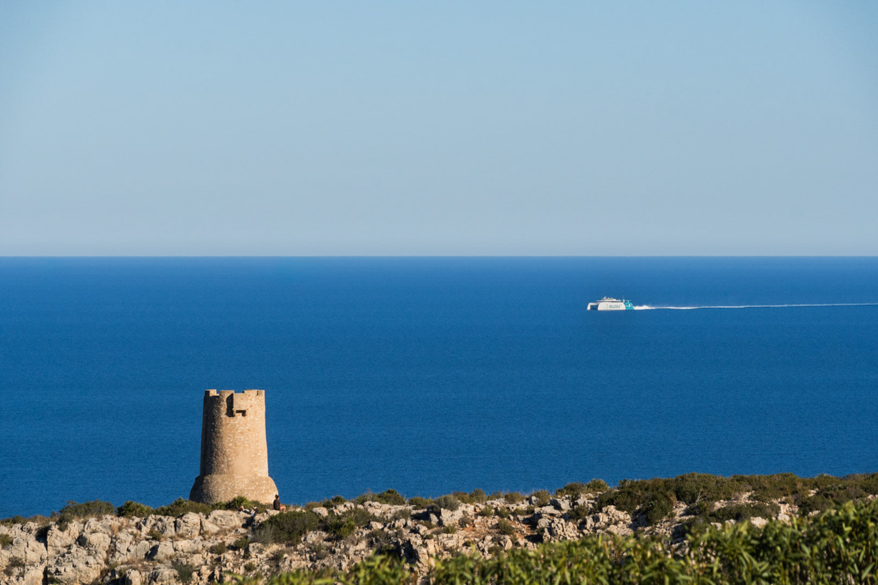 Panoramablick auf die Küste von Las Rotas in Dénia mit dem historischen Wachturm Torre del Gerro und einer Balearia-Fähre auf dem Mittelmeer.