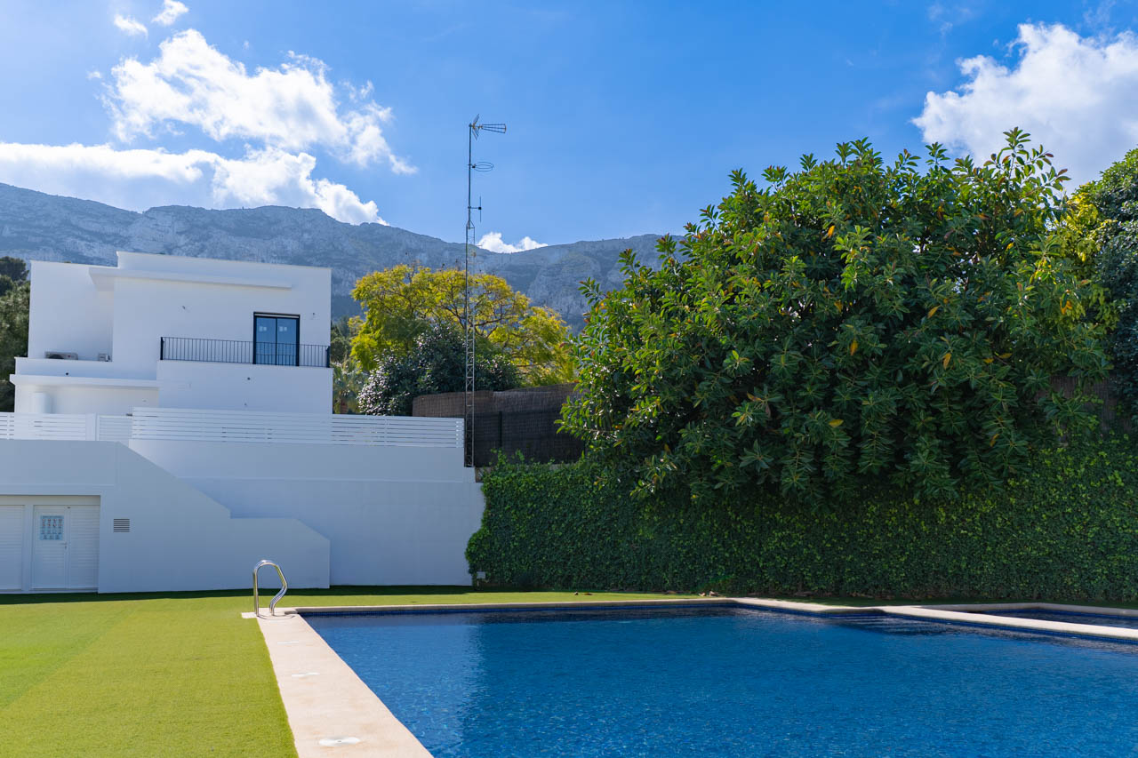 Rechteckiger Pool mit Steineinfassung und Rasenfläche vor einer minimalistischen weißen Fassade mit Blick auf das Montgó-Massiv im Hintergrund.