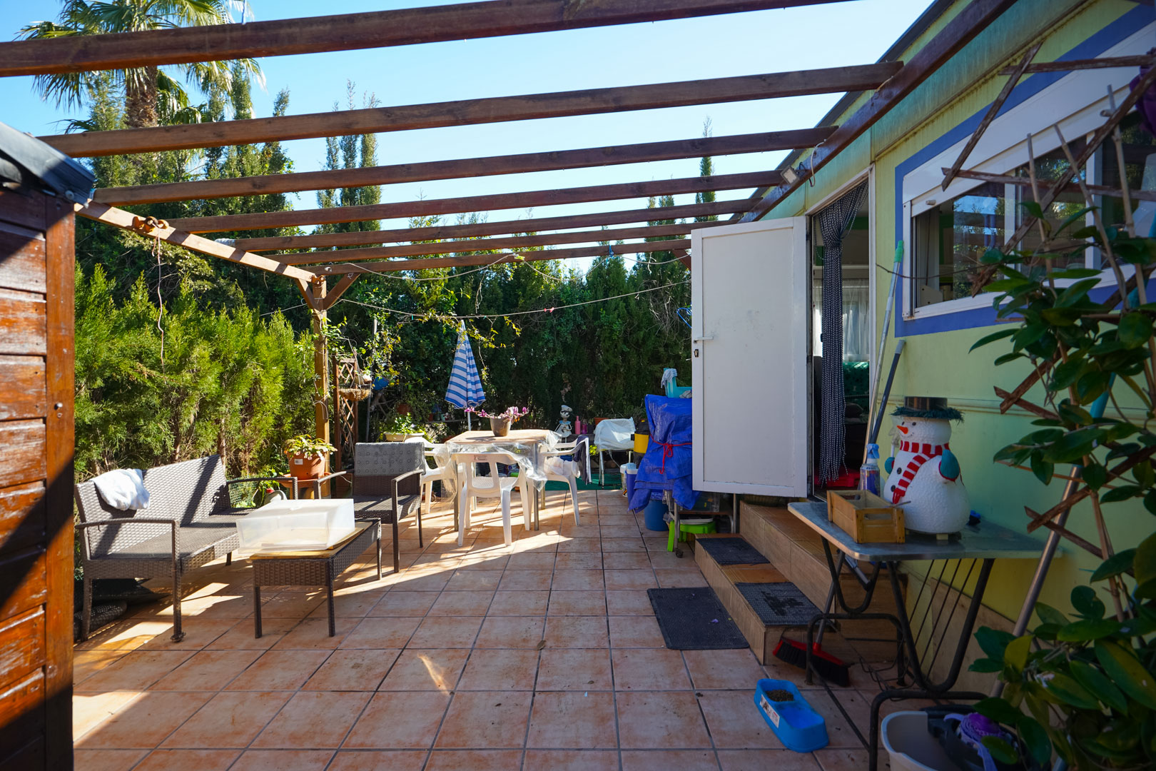 Außen-Terrasse des Fertighauses mit Keramikboden, Holzpergola und Sitzbereich, umgeben von einer blickdichten Hecke.