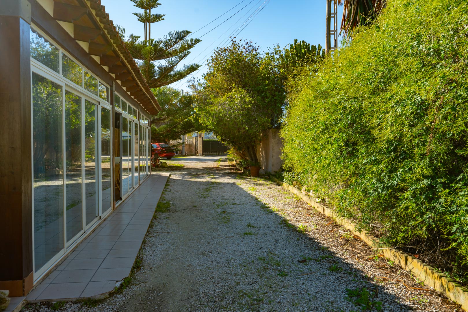 Außenansicht der verglasten Naya mit Holzbalken, gelegen an einem Kiesweg, der von üppiger mediterraner Bepflanzung gesäumt wird.