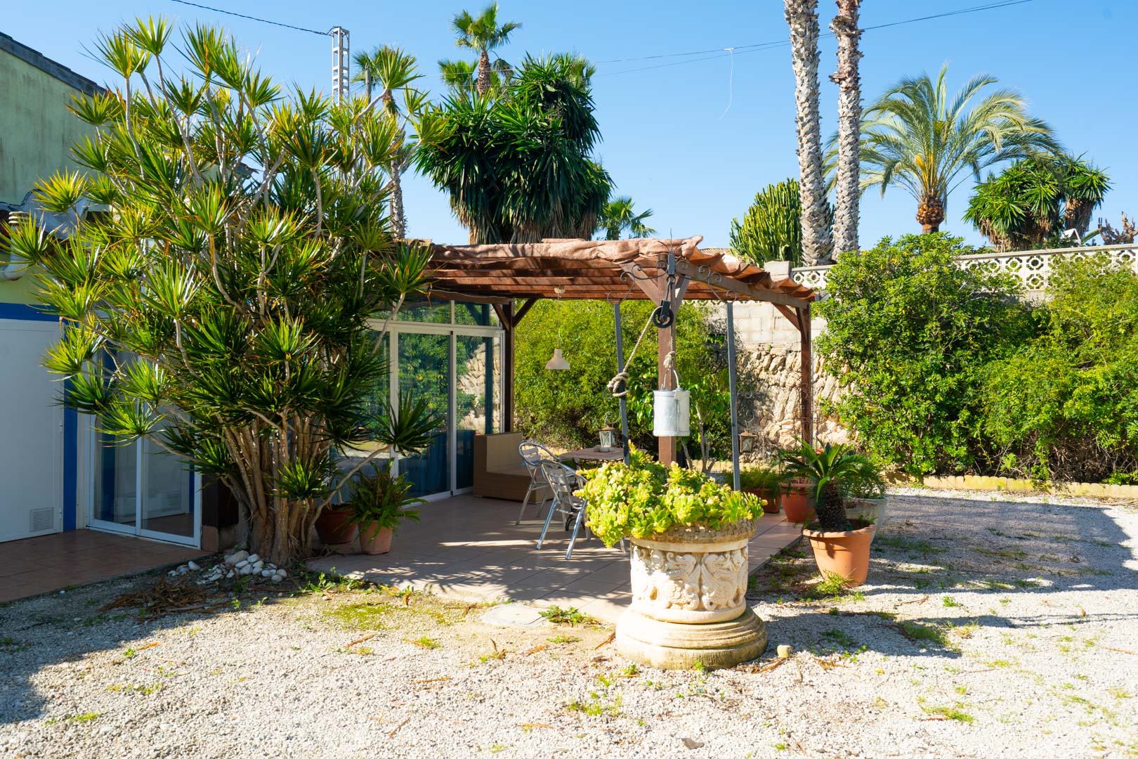 Außenbereich mit Holzpergola, Essplatz und üppiger mediterraner Bepflanzung, ergänzt durch dekorative Steinvasen auf einem Kies- und Fliesenboden.
