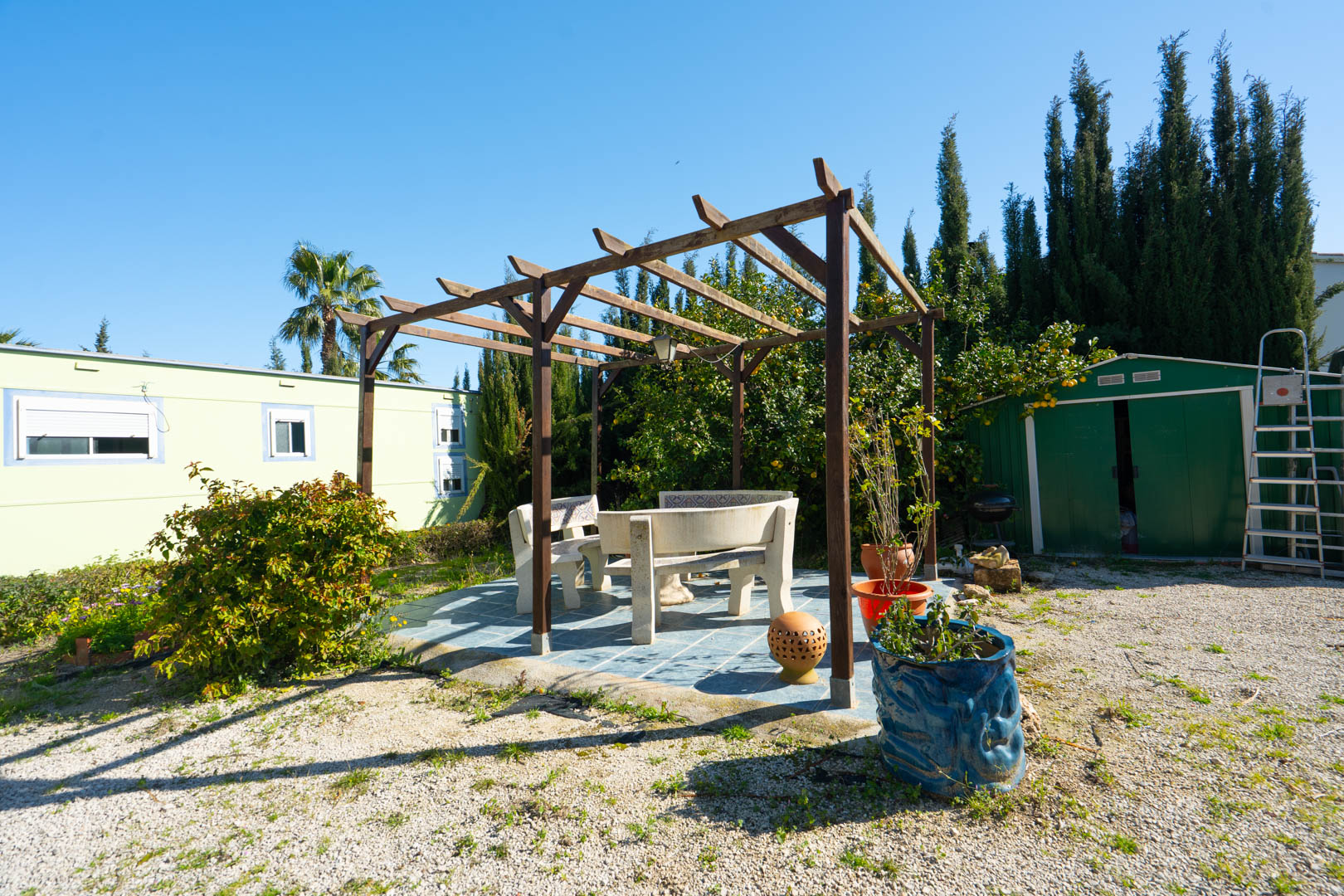 Außenbereich mit Holzpergola und Steinbänken auf Fliesenboden, gelegen zwischen dem Geräteschuppen und dem vorgefertigten Gästehaus.