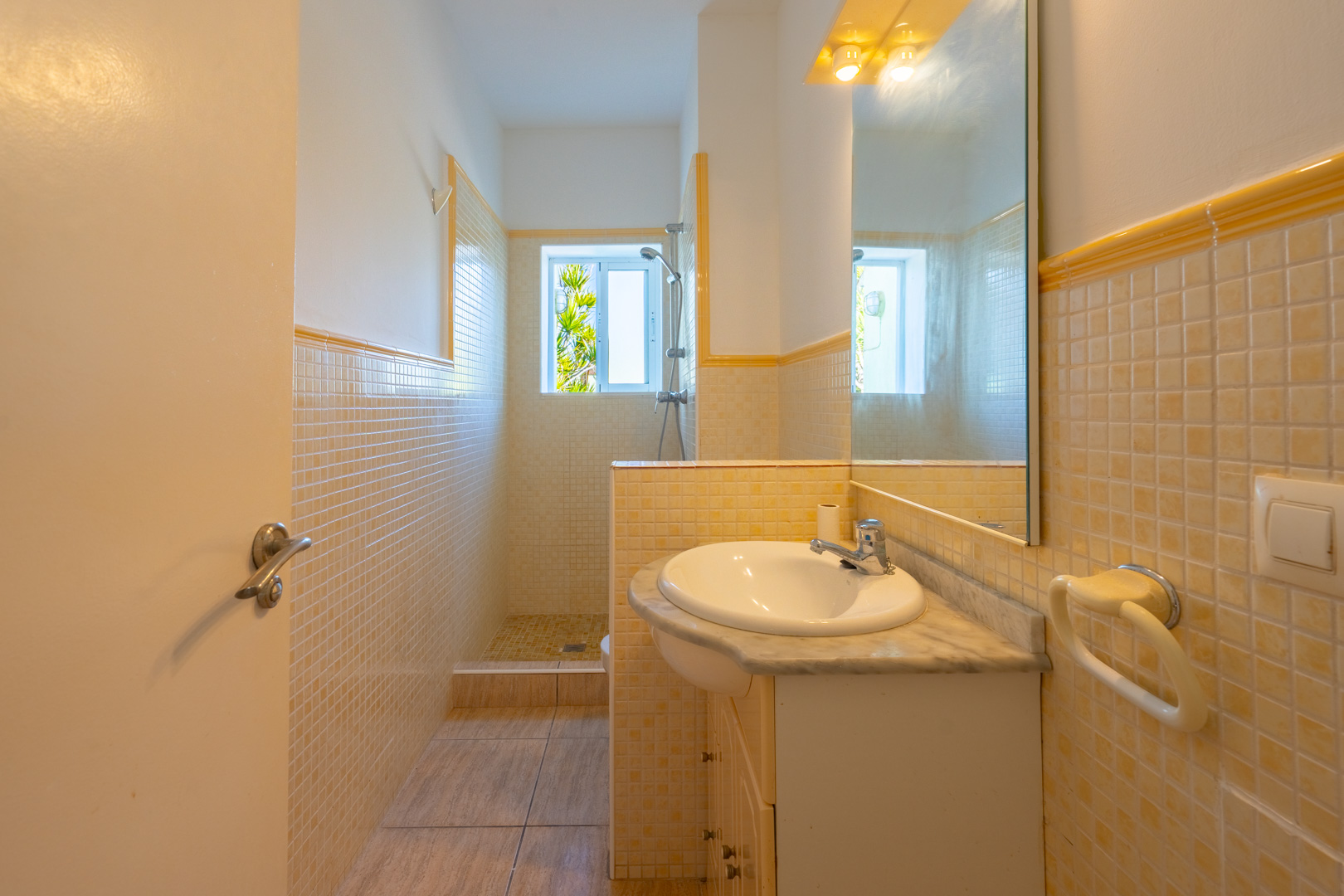 Badezimmer mit begehbarer Dusche, gelb-beigen Mosaikfliesen, einem Waschtisch mit Marmorplatte und Tageslicht durch ein kleines Fenster.