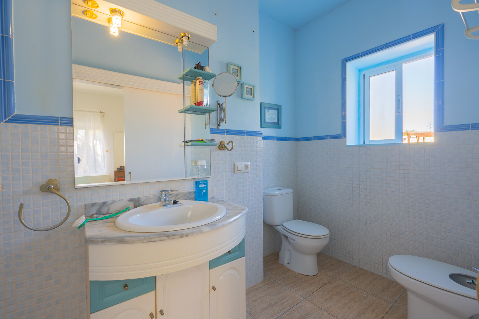 Badezimmer mit blau-weißen Mosaikfliesen, einem Waschtisch mit Marmorplatte und natürlichem Licht durch ein Seitenfenster.