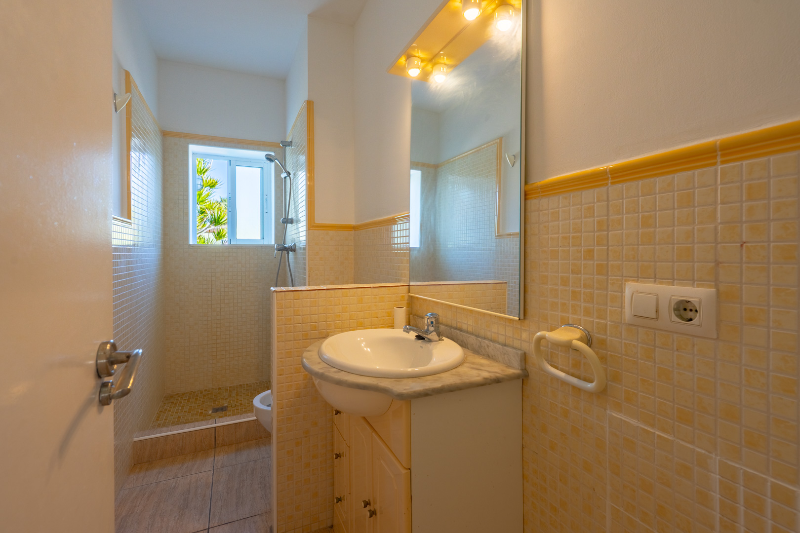Badezimmer mit Mosaik-Dusche, Marmor-Waschtisch und großem Spiegel mit Beleuchtung. Ein Fenster sorgt für natürliches Licht und Ausblick ins Grüne.