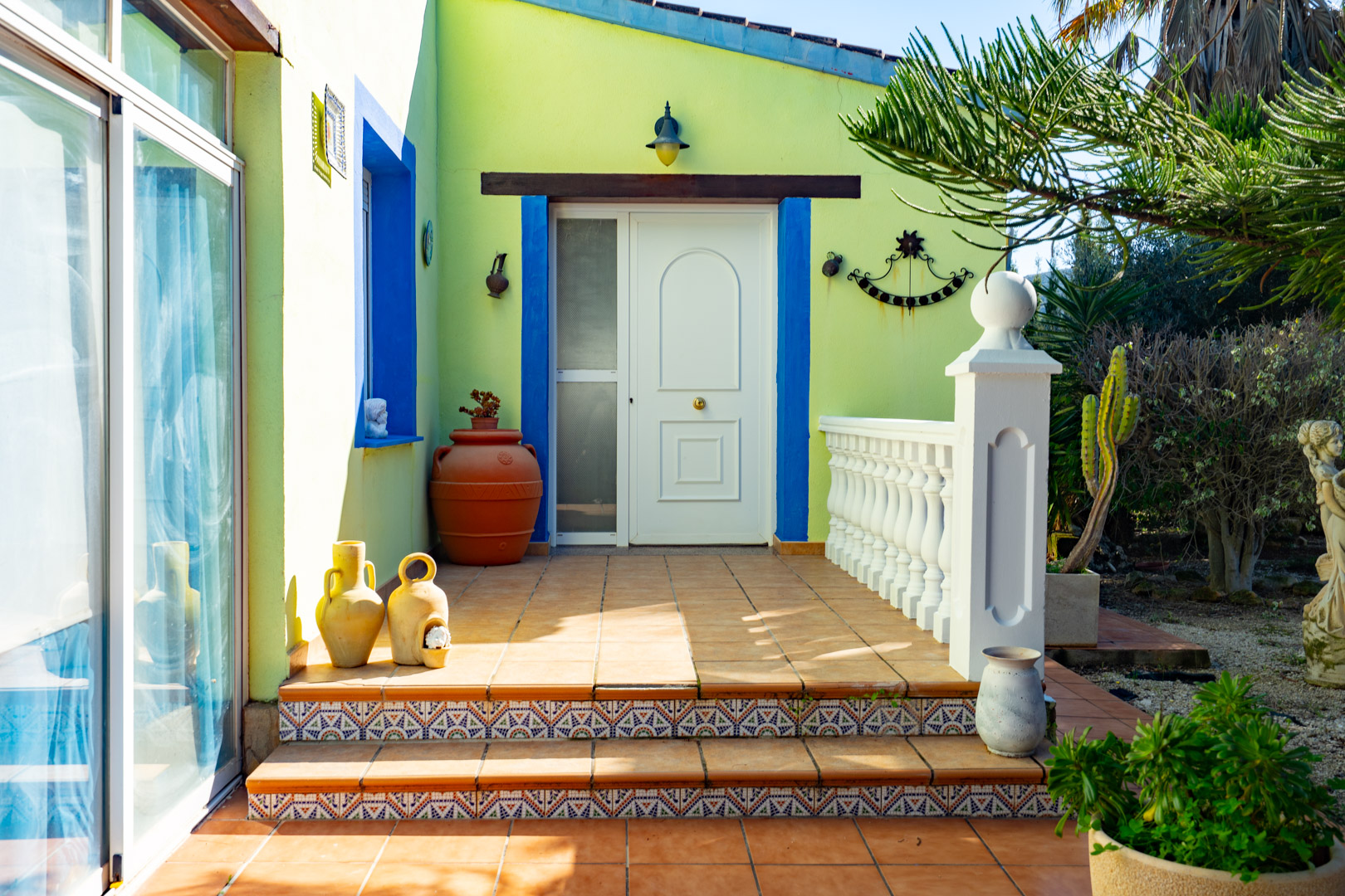 Haupteingang mit weißer Tür, blauen Fensterrahmen und dekorativen Fliesentreppen. Die Terrakotta-Terrasse ist mit einer weißen Balustrade und mediterraner Keramik gesäumt.