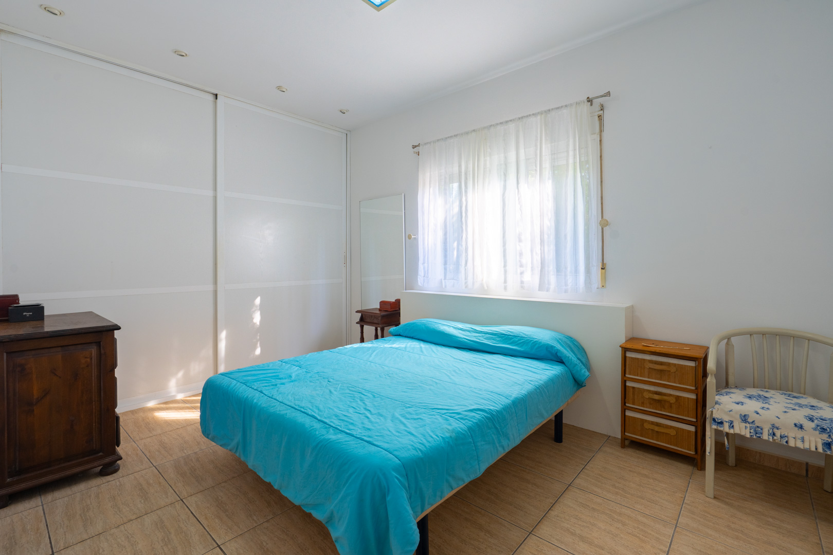 Helles Schlafzimmer mit weißen Einbauschränken, Fliesenboden, Fenster mit lichtdurchlässigen Vorhängen und klassischen Holzmöbeln.