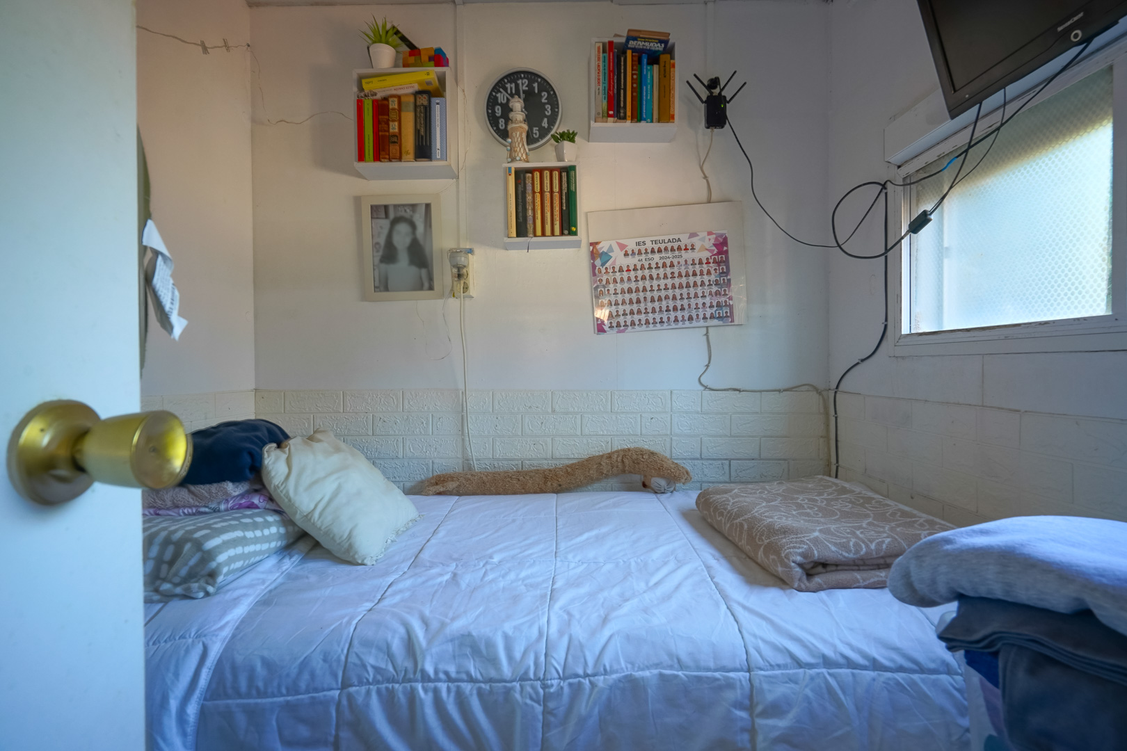 Kompaktes Schlafzimmer mit Einzelbett, Wandregalen und Seitenfenster. Die Wände verfügen über eine dekorative weiße Backstein-Optik im unteren Bereich.