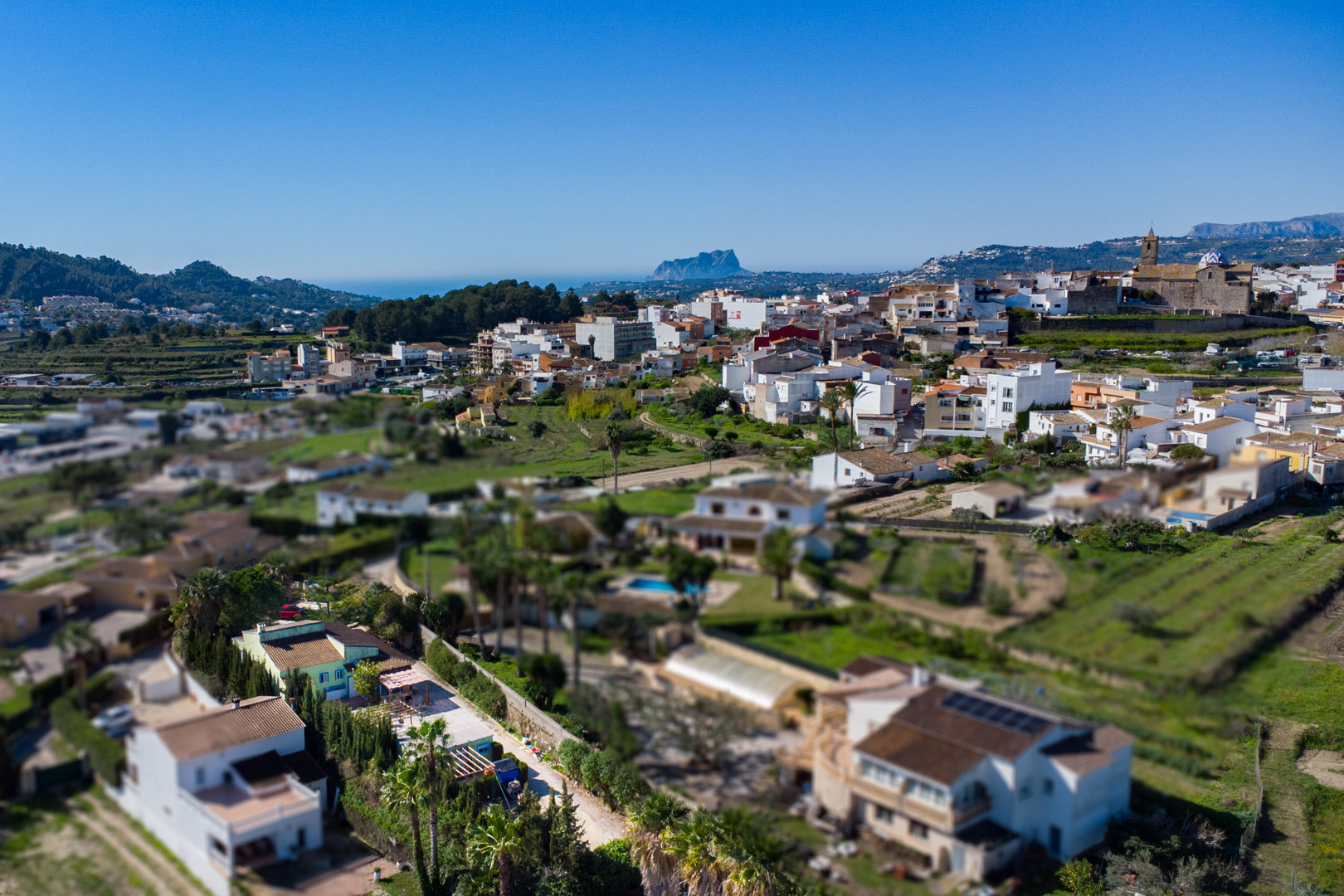 Panorama-Luftaufnahme von Benitachell mit dem Peñón de Ifach am Horizont. Zu sehen sind der Stadtkern, die Pfarrkirche und die mediterrane Landschaft bis zum Meer.