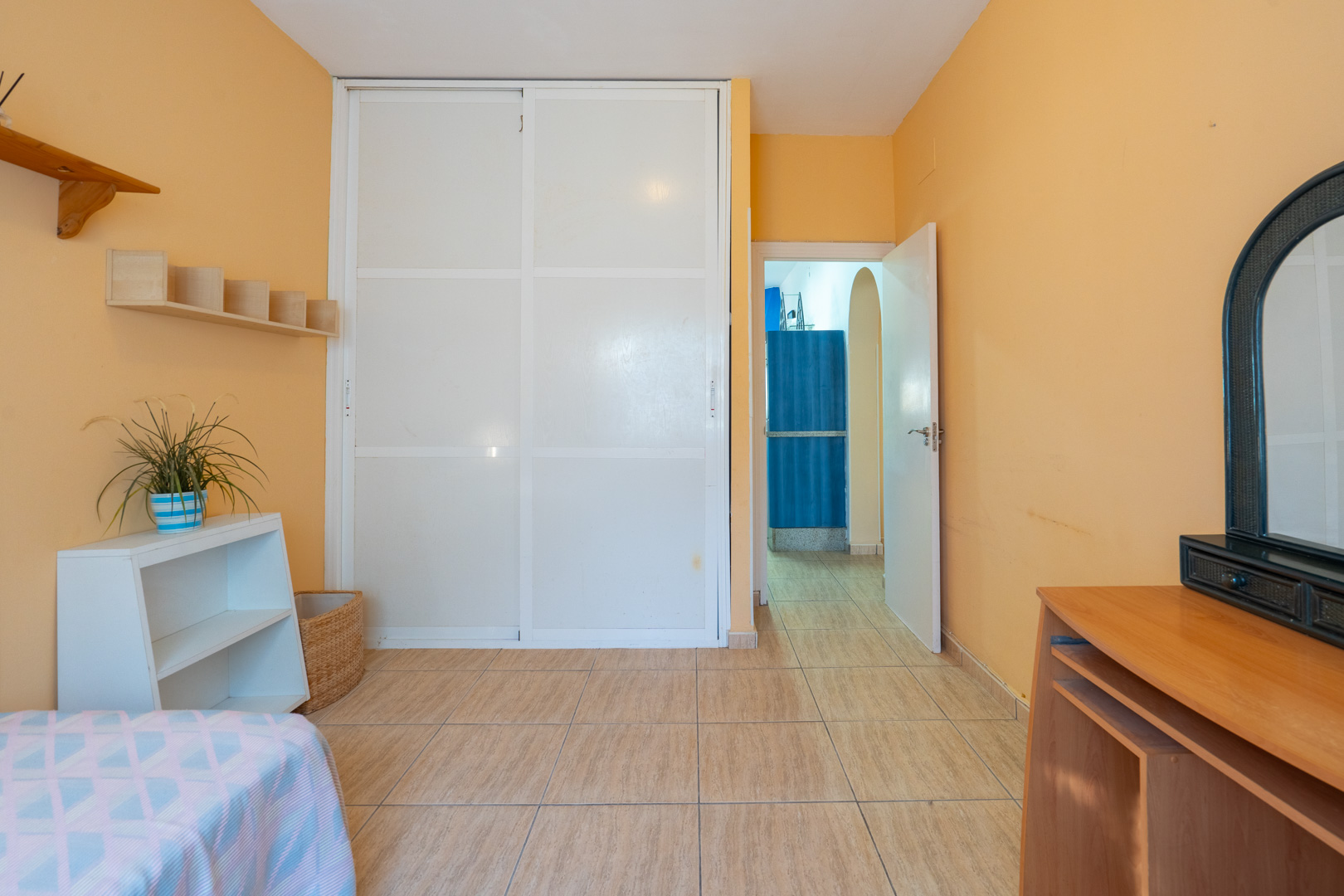 Schlafzimmer mit pfirsichfarbenen Wänden, weißem Einbauschrank mit Schiebetüren, Keramikfliesen und Blick in den angrenzenden Flur.