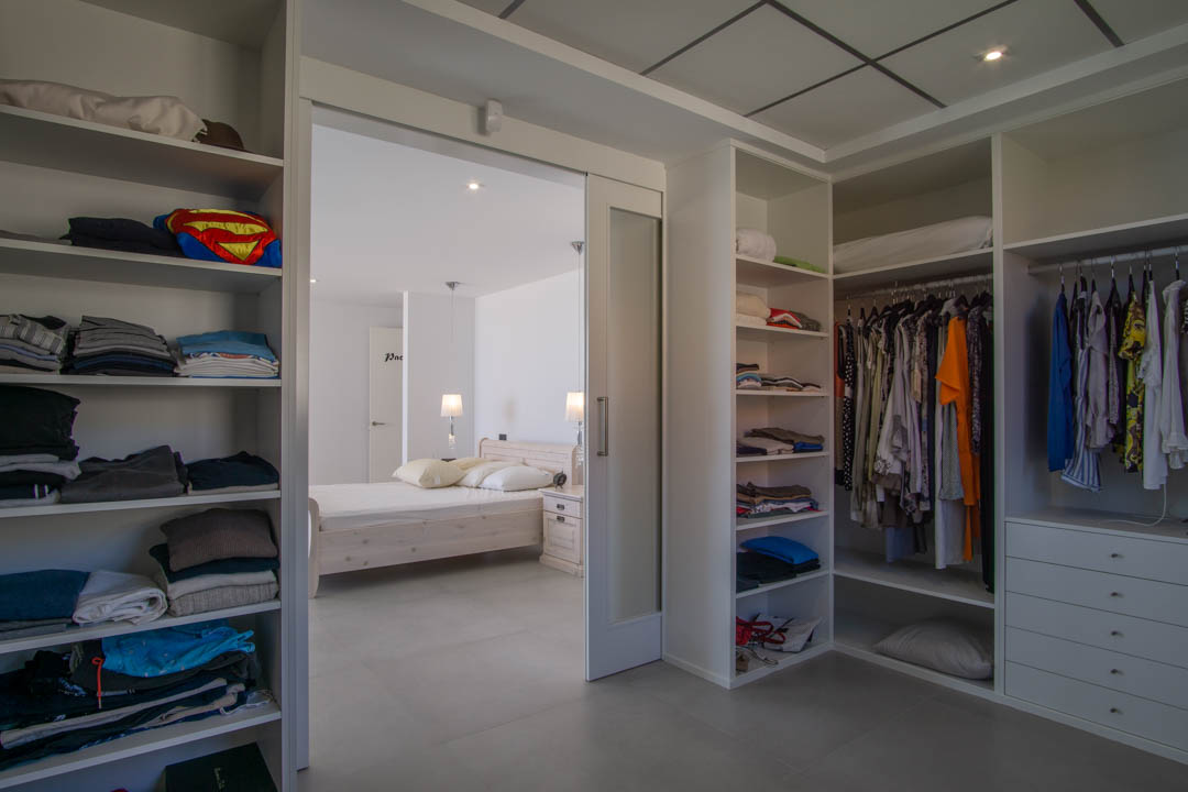 12 m² begehbarer Kleiderschrank mit weißen Regalsystemen, Schubladen und Schiebetür aus Milchglas, die direkt mit dem Hauptschlafzimmer verbunden ist.