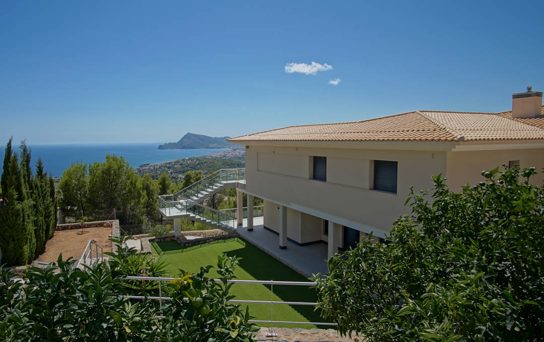 Außenansicht der Villa mit Kunstrasen, Obstbäumen und Glastreppe mit Panoramablick auf die Bucht von Altea und das Mittelmeer.