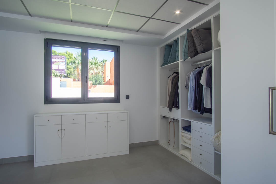 Begehbarer Kleiderschrank mit maßgefertigten weißen Regalsystemen, niedriger Kommode unter dem Fenster und moderner Rasterdecke.