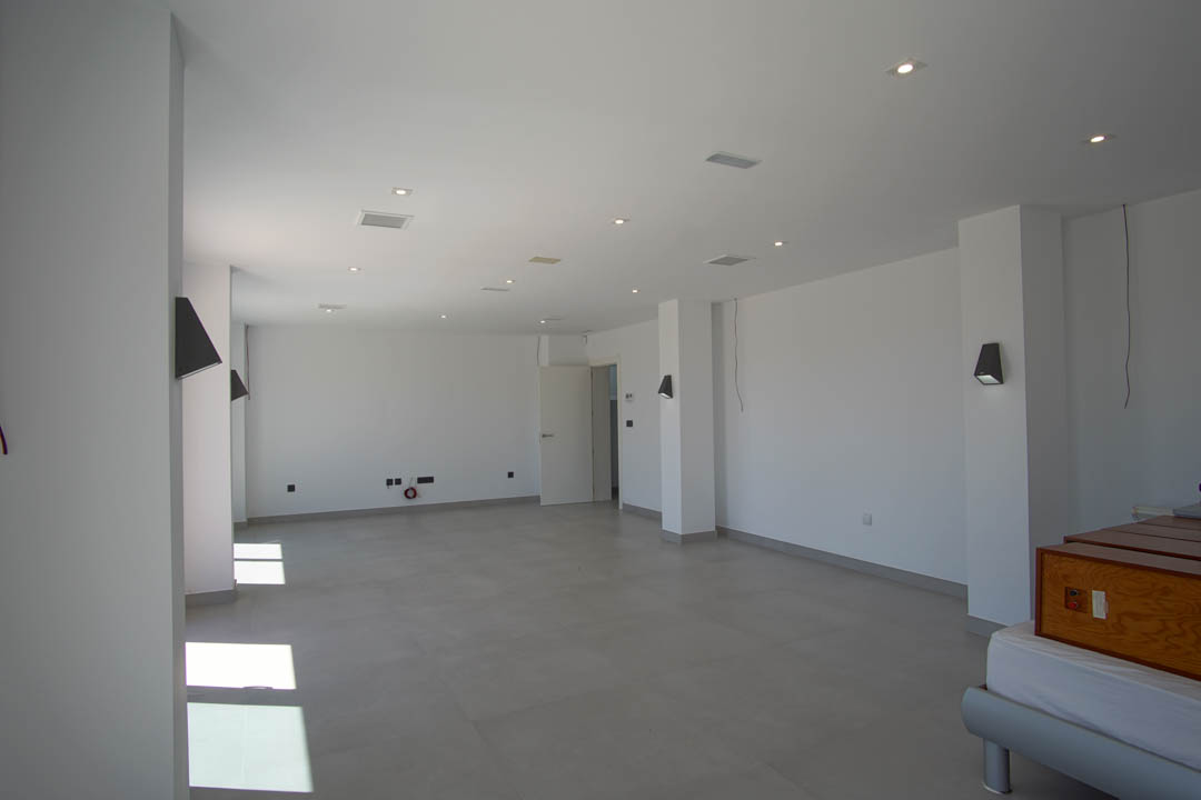 Großzügiger, offener Raum mit grauen Bodenfliesen, weißen Wänden, Deckeneinbaustrahlern und modernen Wandleuchten.