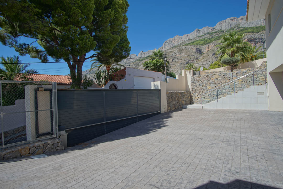 Haupteingang mit großzügiger Einfahrt aus geprägtem Beton, dunklem Schiebetor und Natursteinmauern vor der Kulisse der Sierra de Altea.