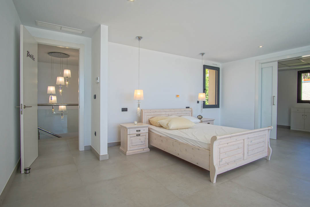 Helles Schlafzimmer mit hellem Holzbett, passenden Nachttischen und Hängeleuchten. Großformatige Fliesenböden und Zugang zum Flur mit modernem Glasgeländer.