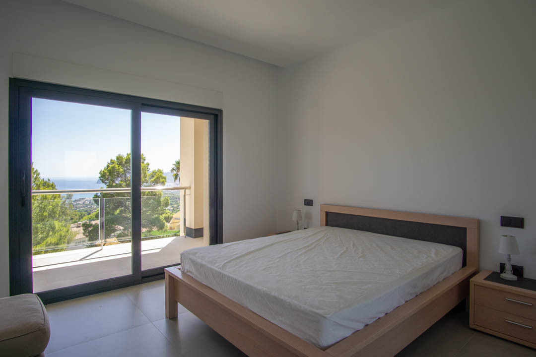 Helles Schlafzimmer mit hellem Holzbettrahmen, großen Glasschiebetüren und privatem Balkon mit Blick auf das Meer und die umliegende Natur.