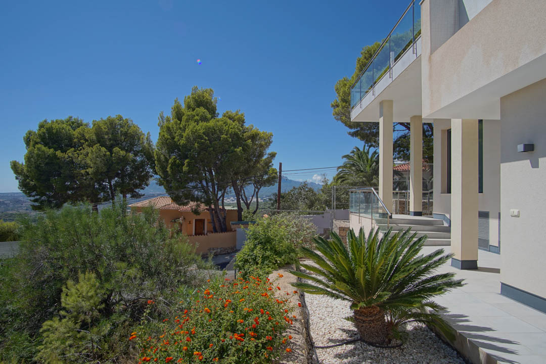 Moderne Außenfassade mit weißen Säulen, Glasgeländern und angelegtem Garten mit Kies, einer Palmfarn und mediterraner Bepflanzung.