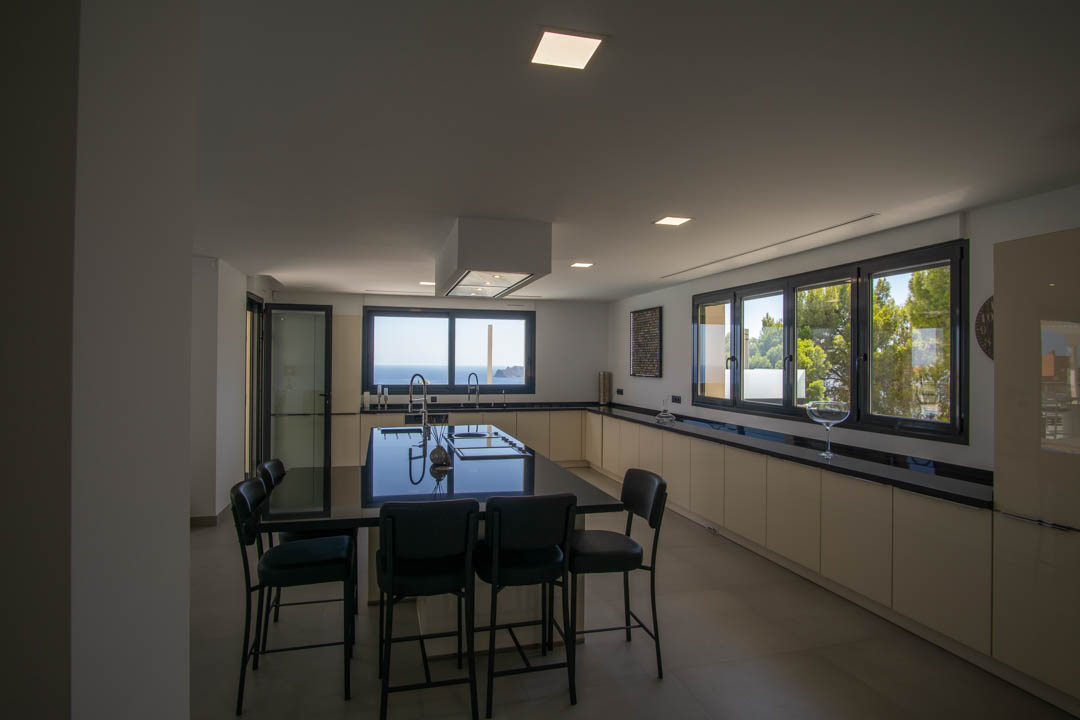 Moderne Küche mit großer schwarzer Granitinsel, minimalistischen cremefarbenen Schränken und großen Fenstern mit Blick auf das Mittelmeer und Pinien.