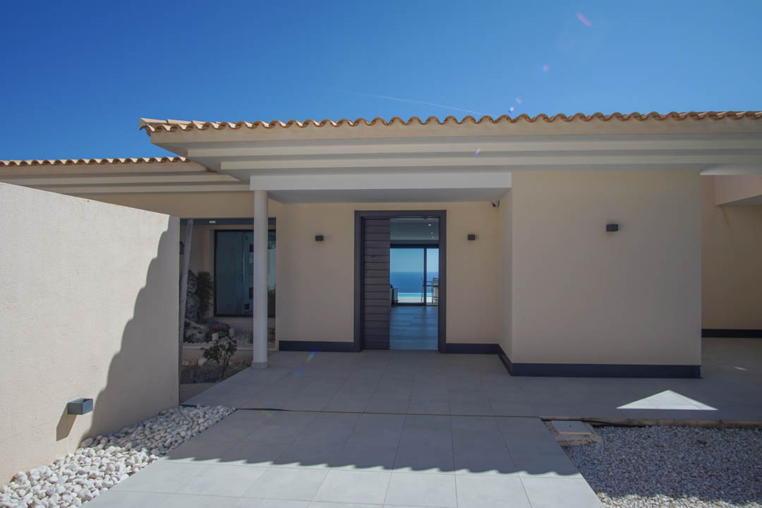 Moderner Haupteingang mit grauer Tür, großformatigen Bodenfliesen und direktem Durchblick durch das Haus auf das azurblaue Mittelmeer.