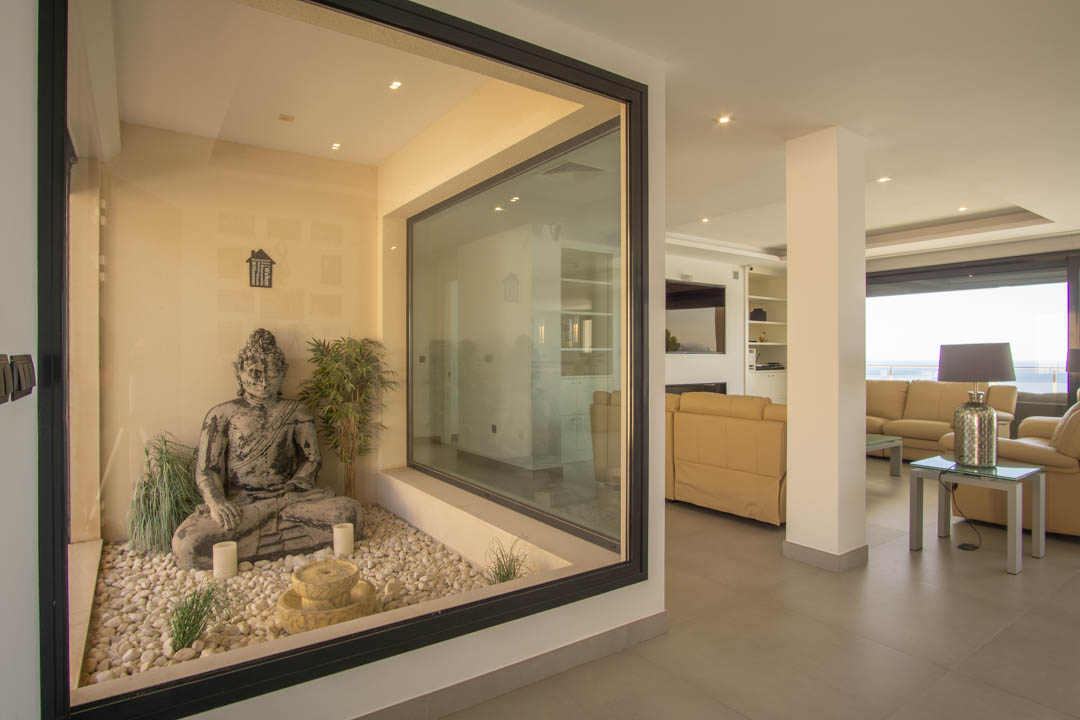 Moderner Wohnbereich mit verglastem Zen-Innengarten, Buddha-Statue und weißen Kieseln, angrenzend an eine Sitzecke mit Ledersofas und Blick auf die Küste.
