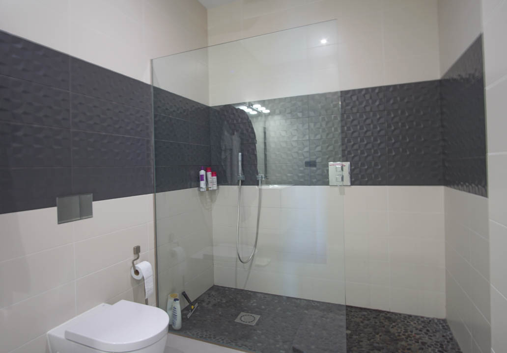 Modernes Badezimmer mit begehbarer Dusche, Glastrennwand, Kieselsteinboden und kontrastierenden Wandfliesen mit geometrischem Relief.