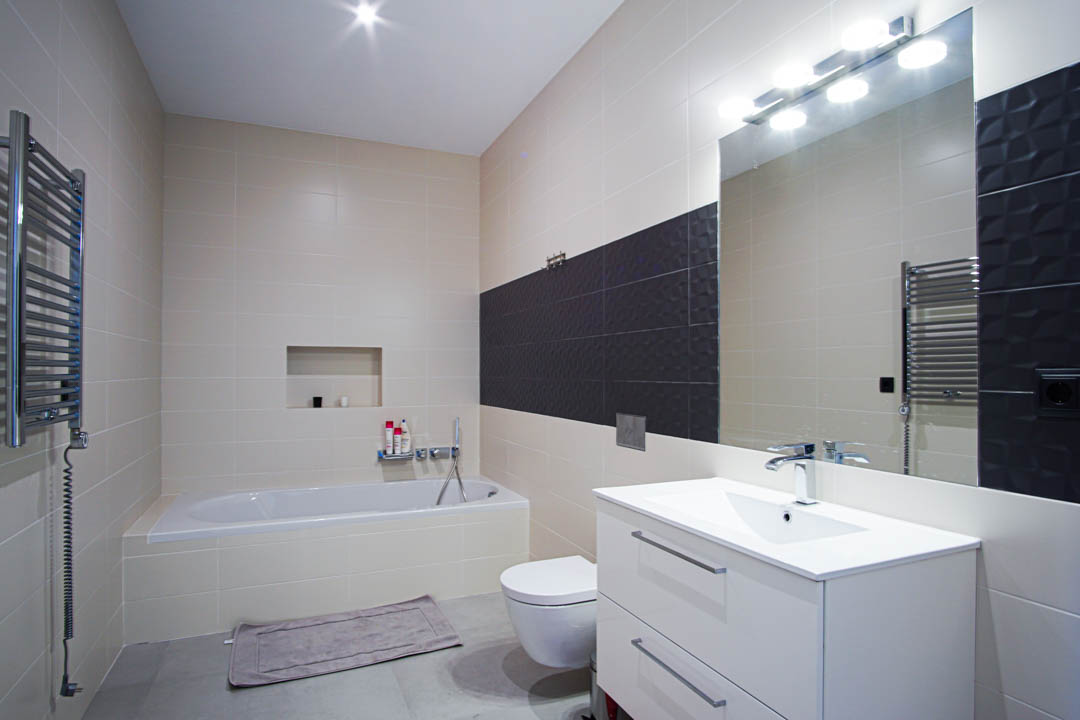 Modernes Badezimmer mit Einbaubadewanne, weißem Waschtisch, wandhängendem WC und beigen Fliesen mit einem dunkelgrauen, texturierten Akzentstreifen.