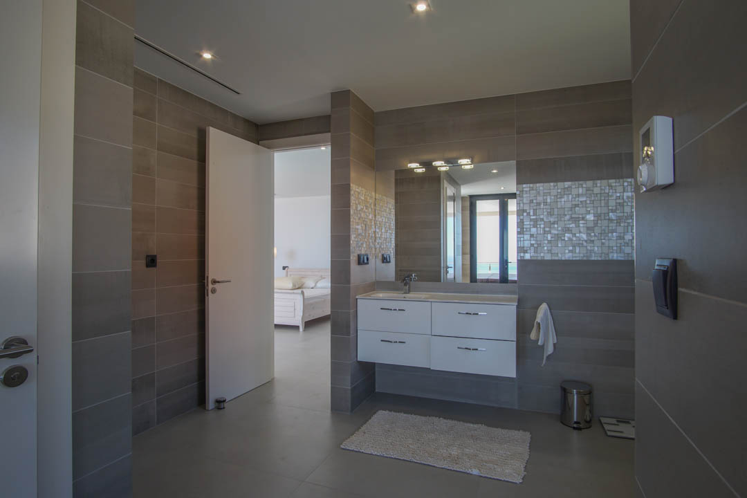 Modernes Badezimmer mit grauen Fliesen, weißem Hängewaschbecken und Mosaik-Akzenten. Die offene Tür führt zum Hauptschlafzimmer mit Blick auf das Mittelmeer.