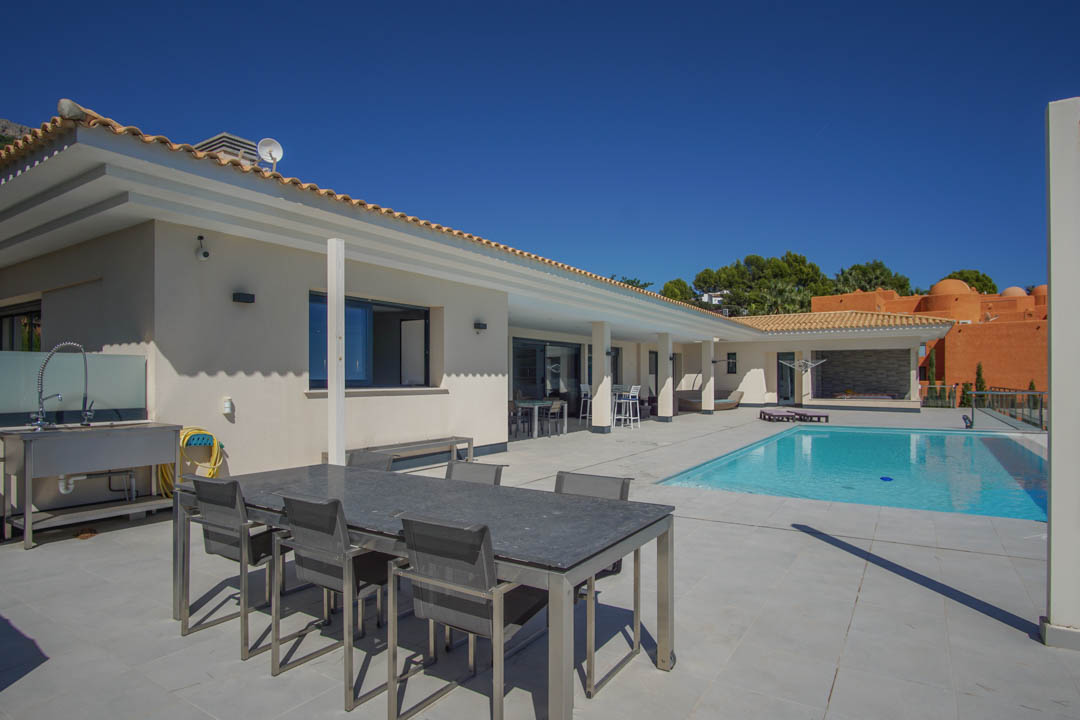 Weitläufige Außenterrasse mit rechteckigem Pool, Essbereich mit Steintisch und industrieller Edelstahlspüle vor der weißen Fassade der Villa.
