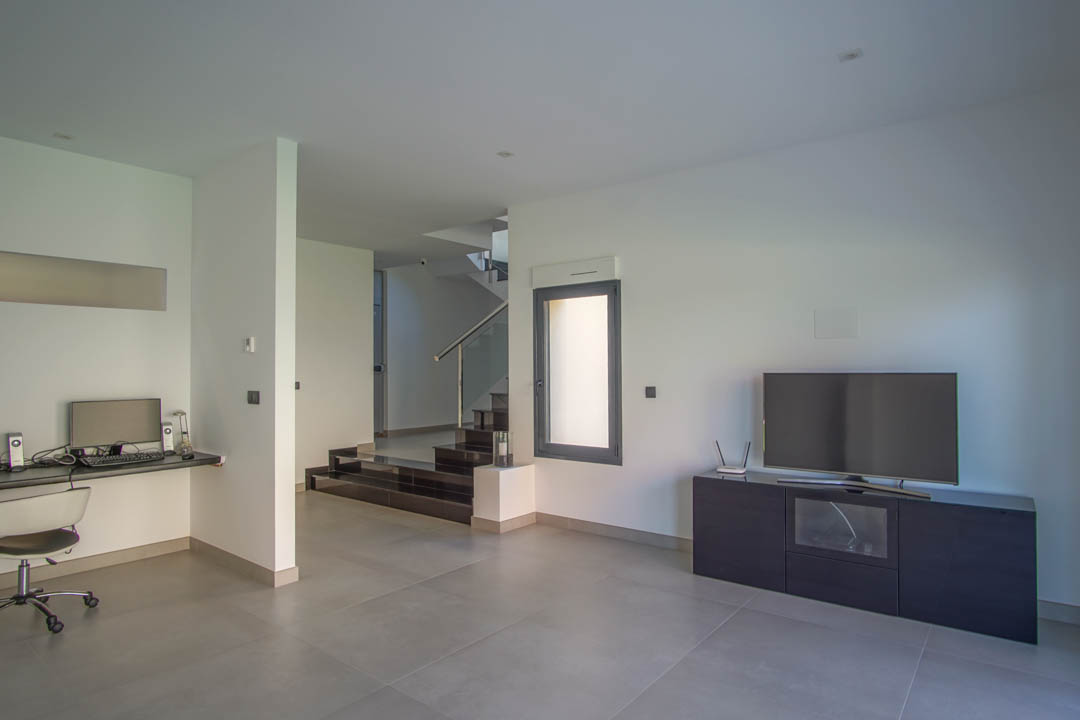 Wohnzimmer im Erdgeschoss mit integriertem Arbeitsbereich, minimalistischem TV-Möbel und Treppe mit dunklen Stufen und Glasgeländer.