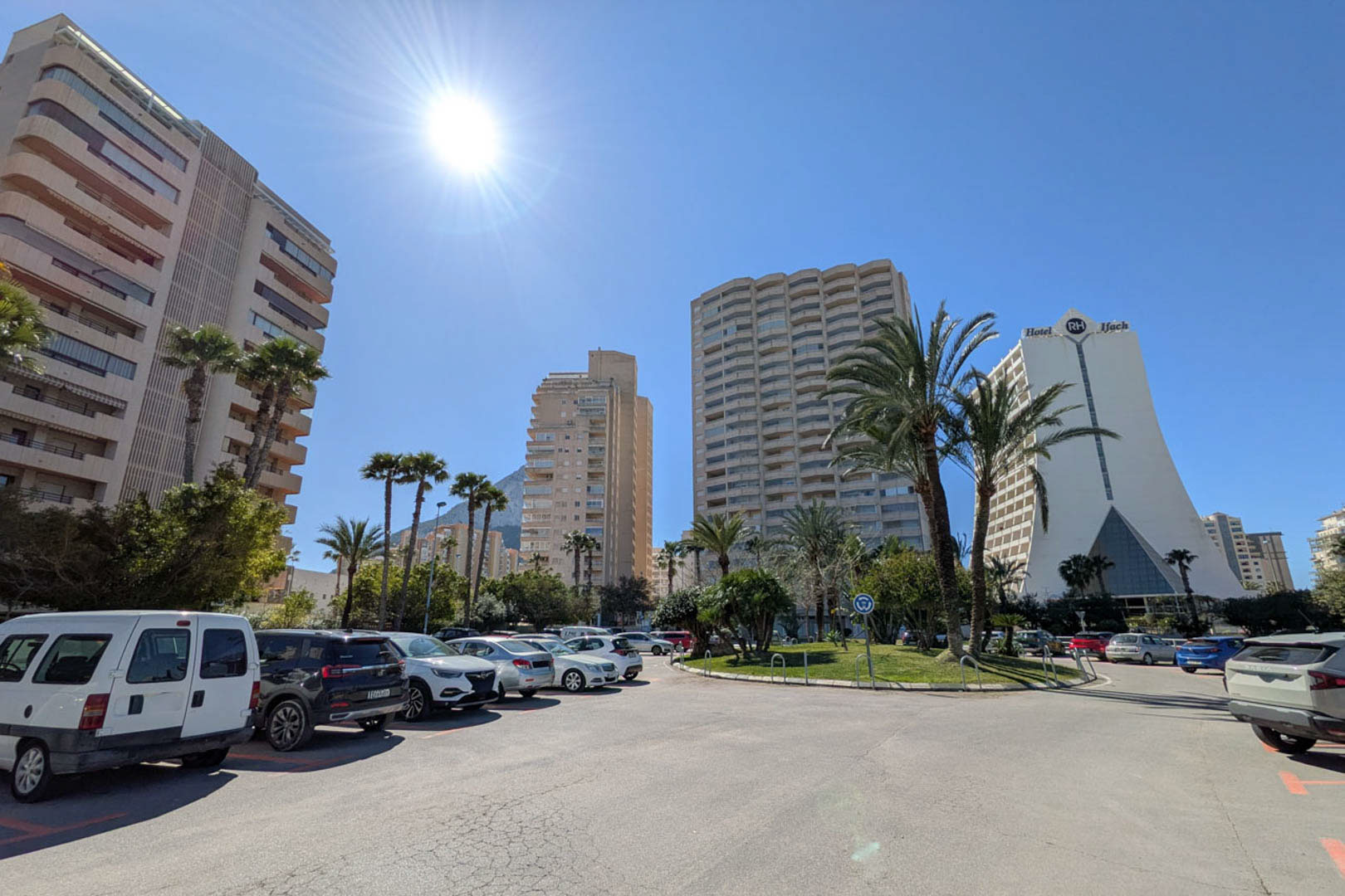 Außenansicht eines Stadtviertels in Calpe mit markanten Wohnhochhäusern, dem Hotel RH Ifach und öffentlichen Parkplätzen bei strahlendem Sonnenschein.
