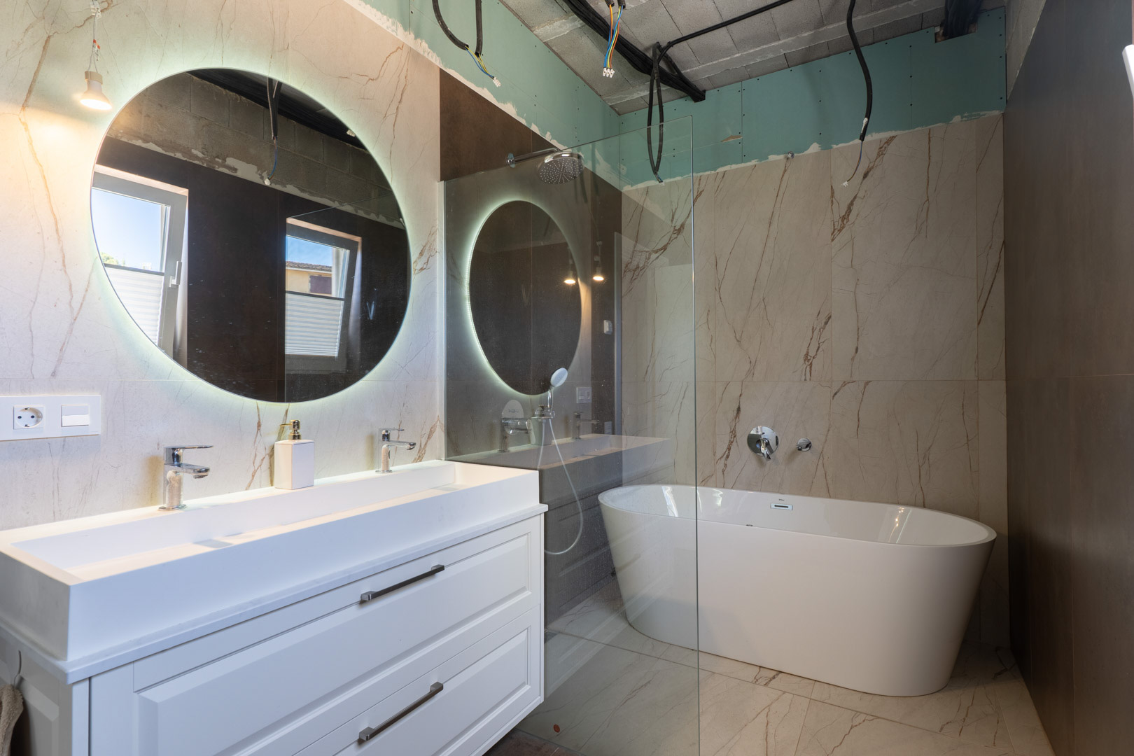 Badezimmer im Bau mit Doppelwaschtisch, hinterleuchtetem Rundspiegel und freistehender Badewanne vor großformatigen Feinsteinzeugfliesen.