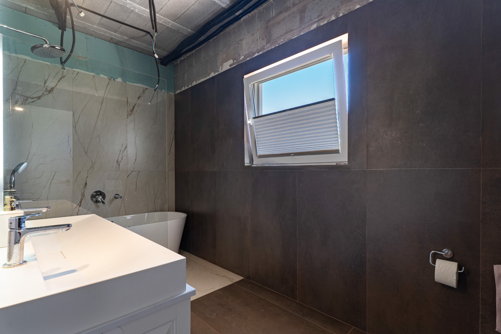 Badezimmer in der Fertigstellungsphase mit Doppelwaschbecken, freistehender Badewanne und Dusche in Marmoroptik. Großformatige dunkle Wandfliesen treffen auf ein praktisches Kippfenster.
