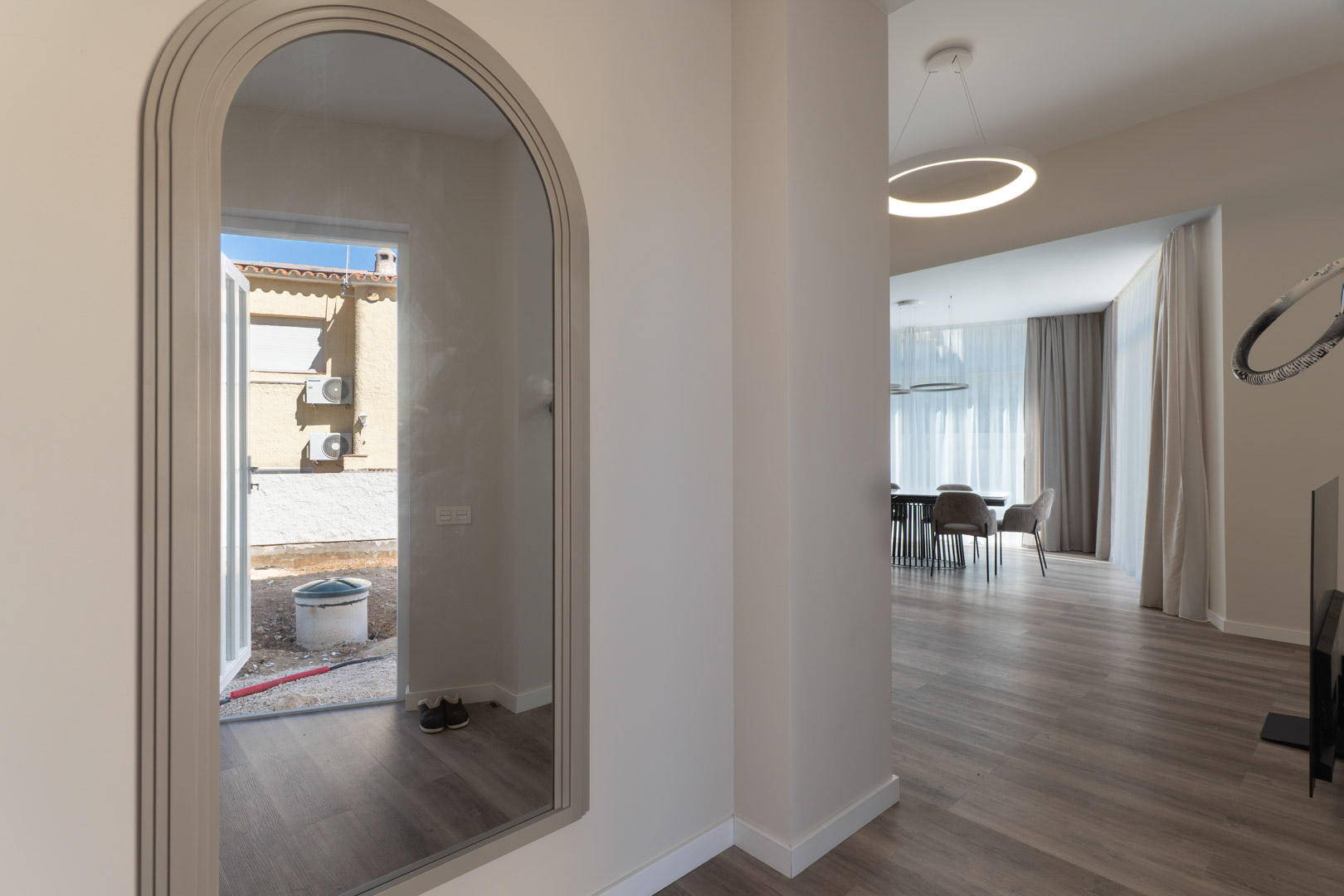 Eingangsbereich mit Rundbogenspiegel und Blick auf den minimalistischen Wohnbereich mit hellem Boden und einer modernen kreisförmigen LED-Leuchte.