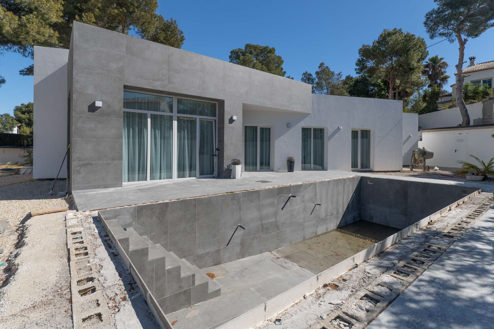 Einstöckige minimalistische Villa mit grauer Feinsteinzeug-Fassade und einem im Bau befindlichen Pool, umgeben von mediterranen Pinien.