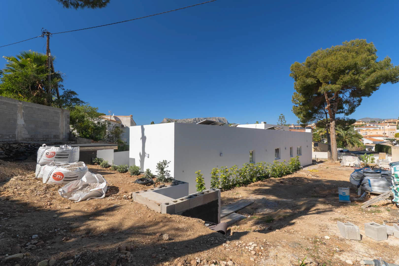 Einstöckige minimalistische Villa mit weißer Fassade und Solarmodulen auf dem Dach. Das Außengelände befindet sich derzeit in der Erschließungs- und Gartenbauphase.