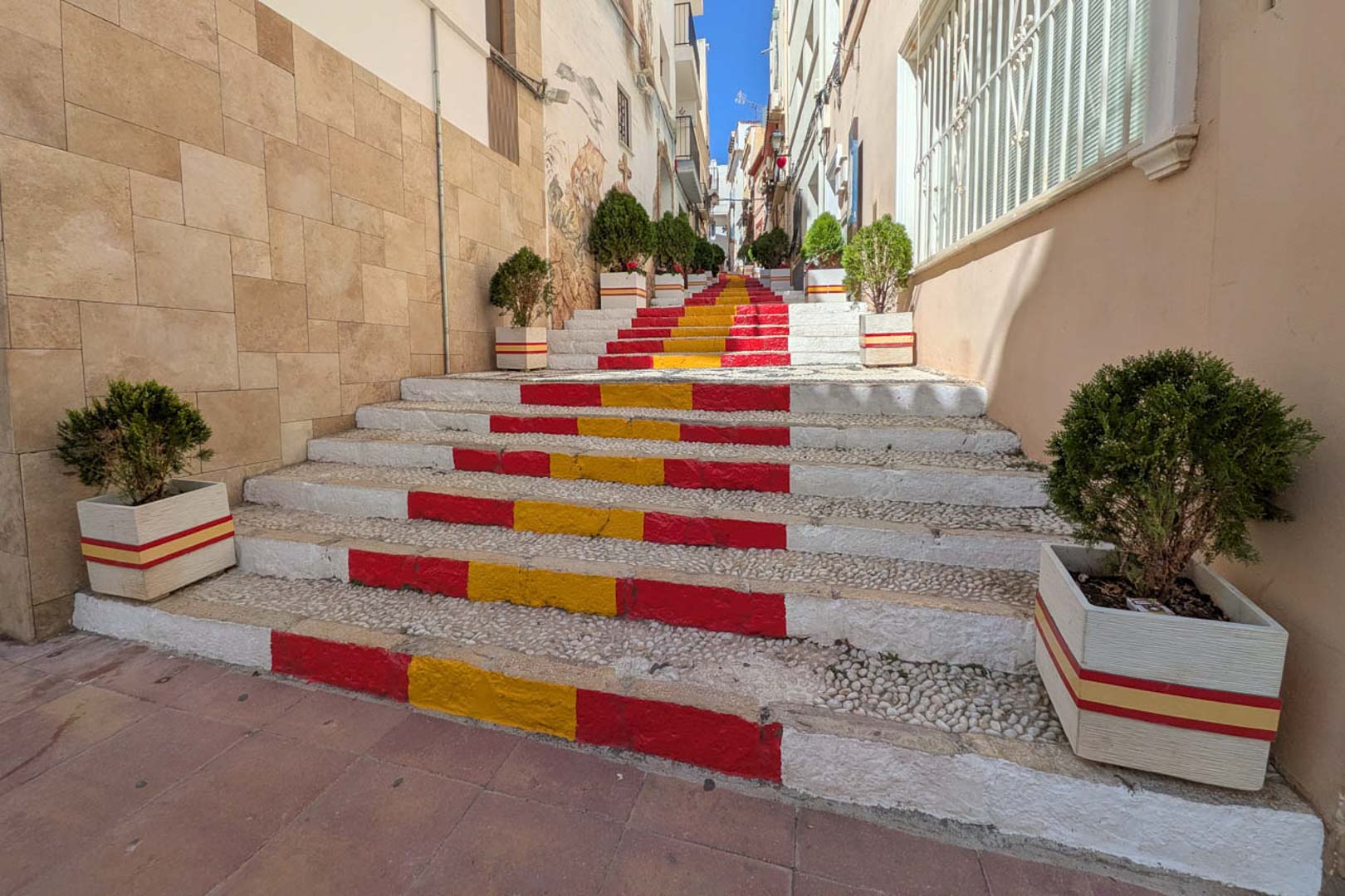 Fußgängerzone in der Altstadt von Calpe mit einer Steintreppe in den spanischen Nationalfarben, gesäumt von dekorativen Pflanzgefäßen und traditionellen Mauern.