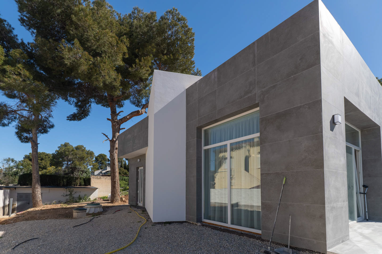Minimalistische Fassade mit grauen Keramikfliesen und großen Glasfronten, eingebettet in ein Grundstück mit altem Baumbestand und unfertigen Außenanlagen.