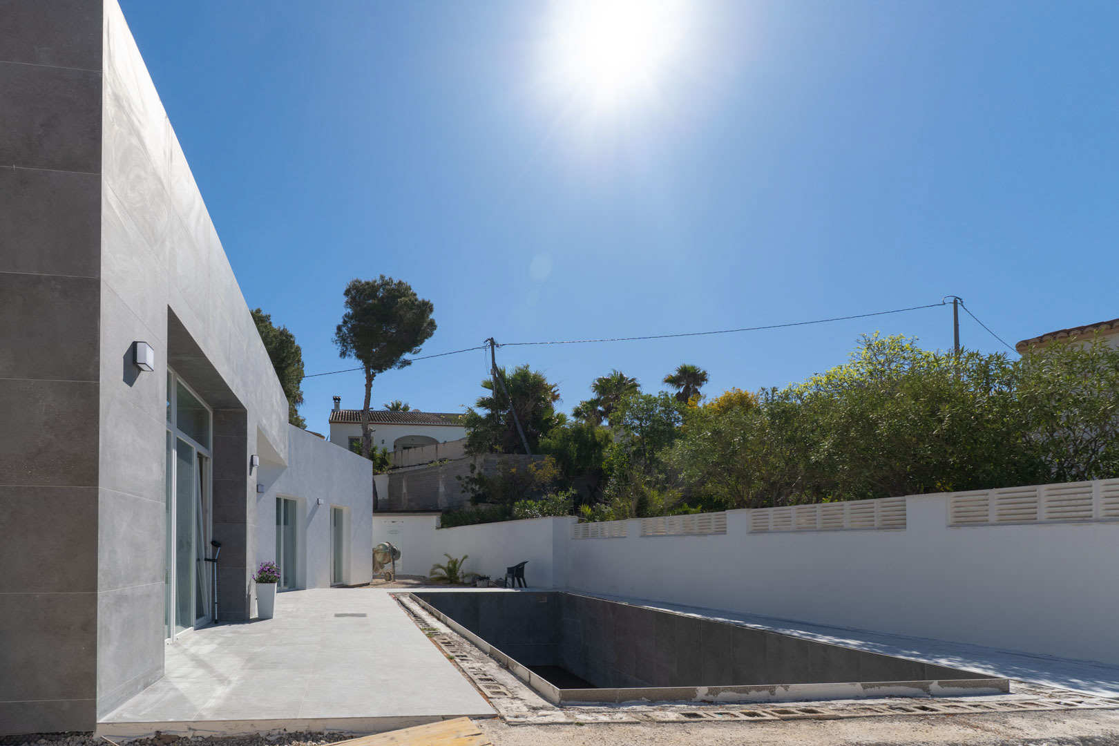 Minimalistische Fassade mit grauer Steinverkleidung und ein im Bau befindlicher Poolbereich, eingefasst von weißen Mauern unter strahlender Sonne.