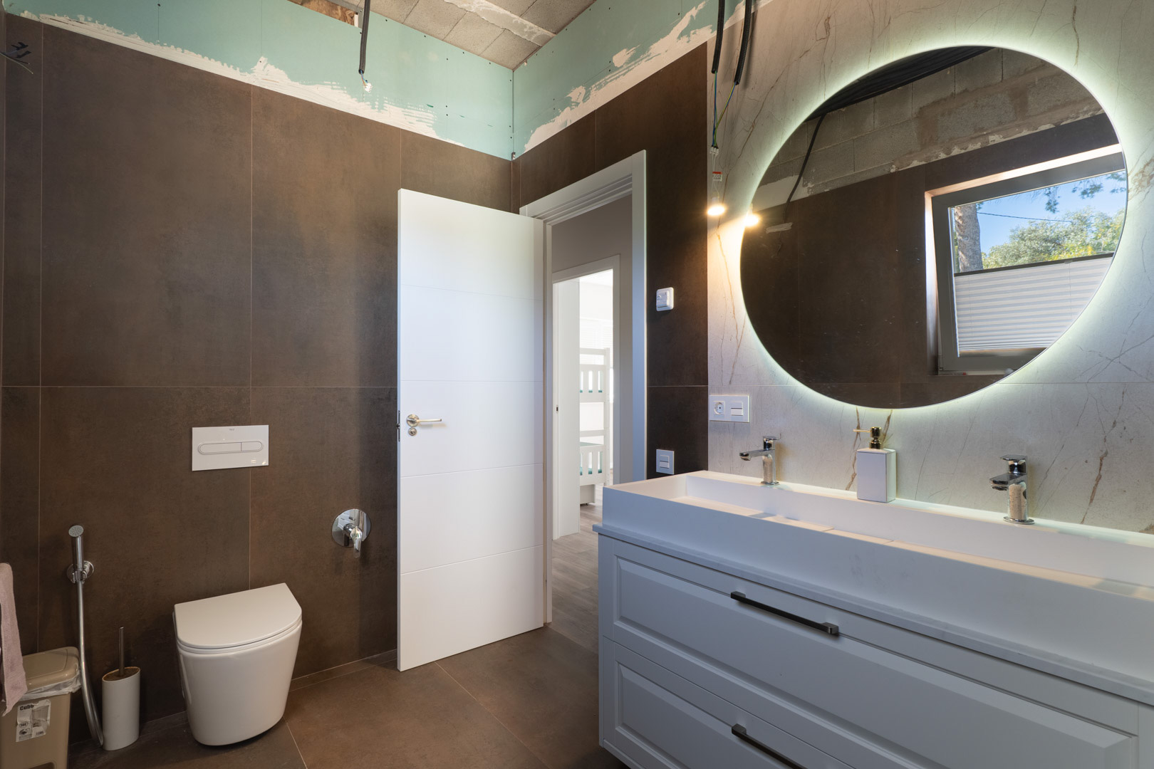 Modernes Badezimmer mit Doppelwaschtisch, hinterleuchtetem Rundspiegel und dunklen Erdtönen. Ausgestattet mit einem Wand-WC und weißem Unterschrank.
