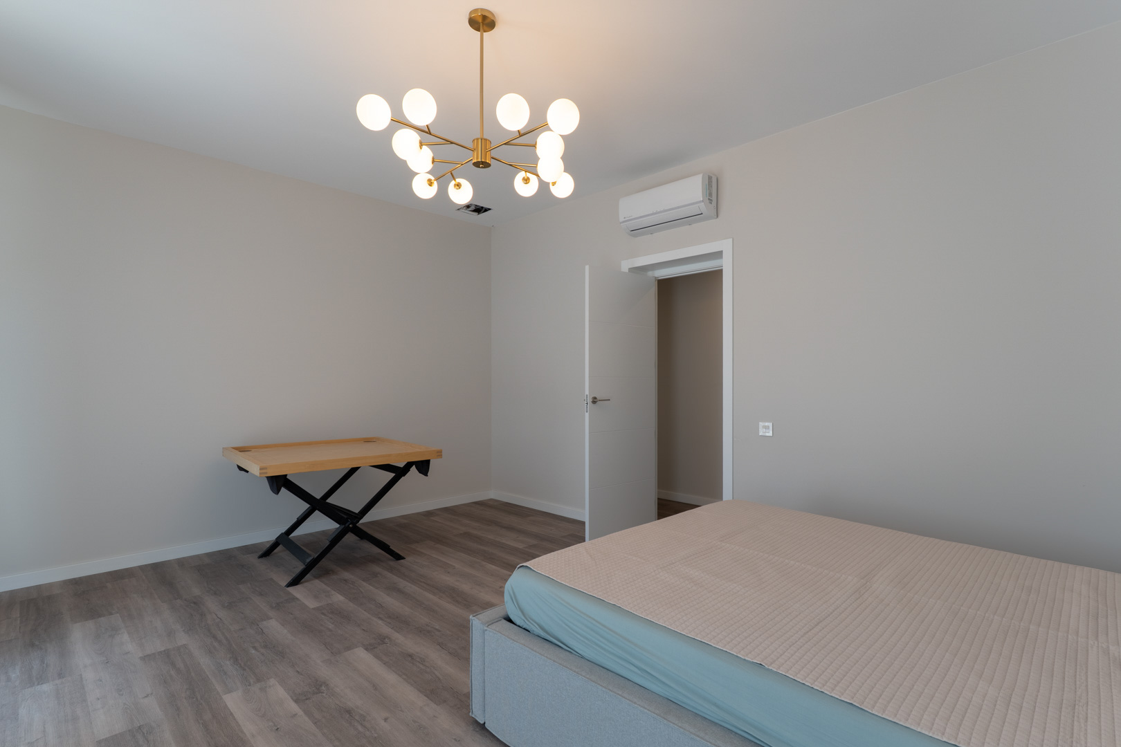 Modernes Schlafzimmer mit grauem Laminatboden, neutralen Wänden und einem goldenen Designer-Kronleuchter. Inklusive Klimaanlage und schlichter weißer Tür.