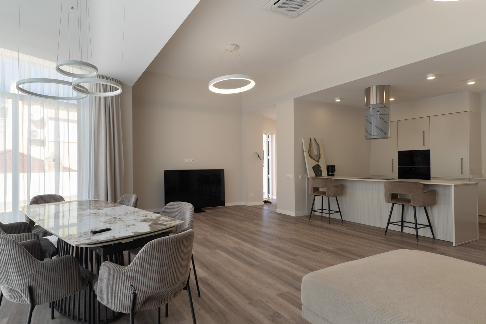 Offener Wohnbereich mit Marmor-Esstisch, minimalistischer Küche samt Frühstücksbar und modernen ringförmigen LED-Pendelleuchten auf Laminatboden.
